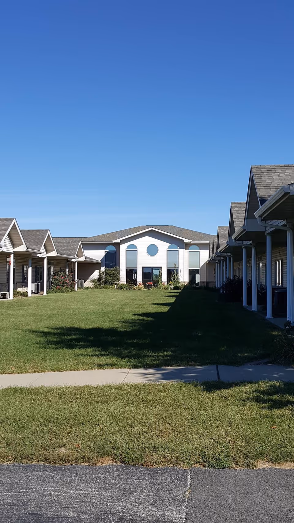 View of a retirement community with a large green lawn in the center, flanked by single-story buildings with covered porches on both sides, and a larger building with multiple windows at the far end under a clear blue sky.