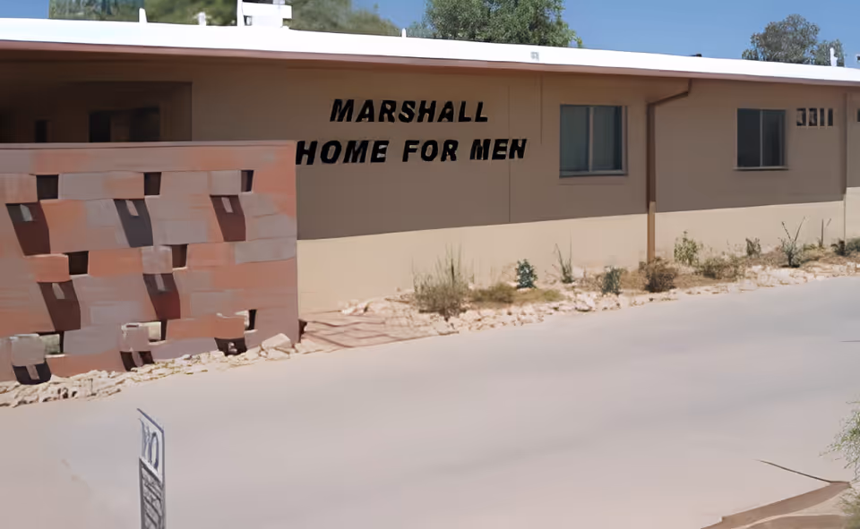 Single-story building exterior with 'Marshall Home for Men' painted on the wall, a decorative block screen, and sparse desert landscaping.