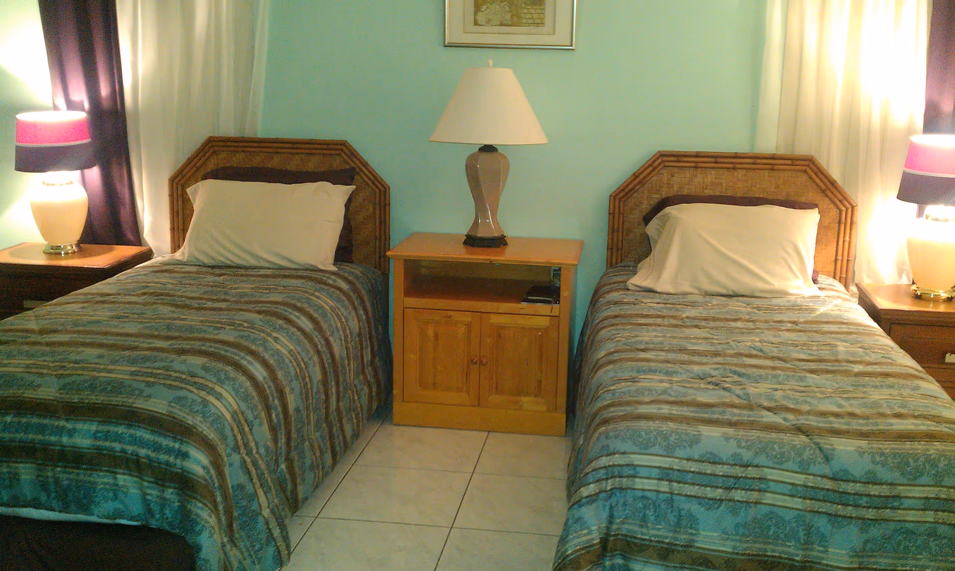 A bedroom with two single beds separated by a wooden nightstand with a lamp on it. Each bed has a pillow and a striped bedspread. There are matching lamps on wooden side tables next to each bed, and the walls are painted light blue with curtains on the windows.