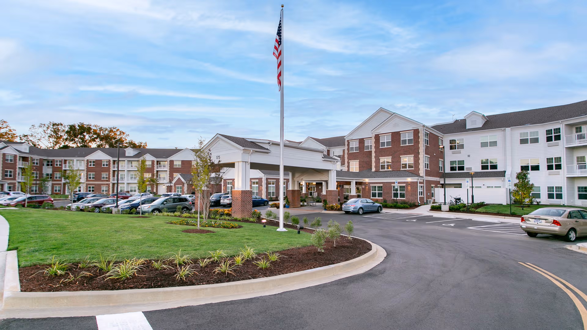 Exterior view of Sycamore Ridge Gracious Retirement Living with a curved drive, flagpole, and parked cars.