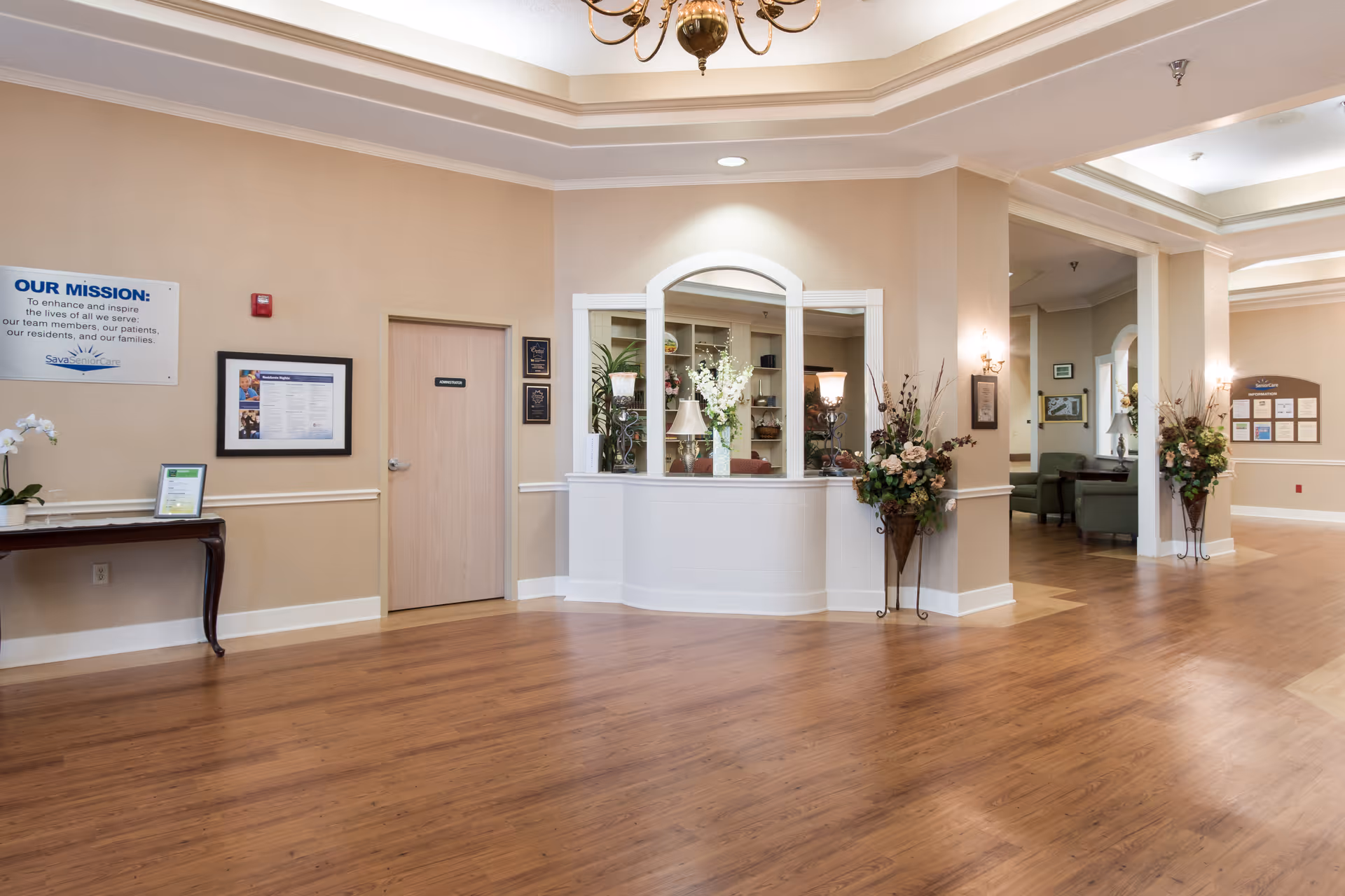 Bright nursing home reception lobby with a white check-in desk, decorative flower arrangements, seating area and wood floors.