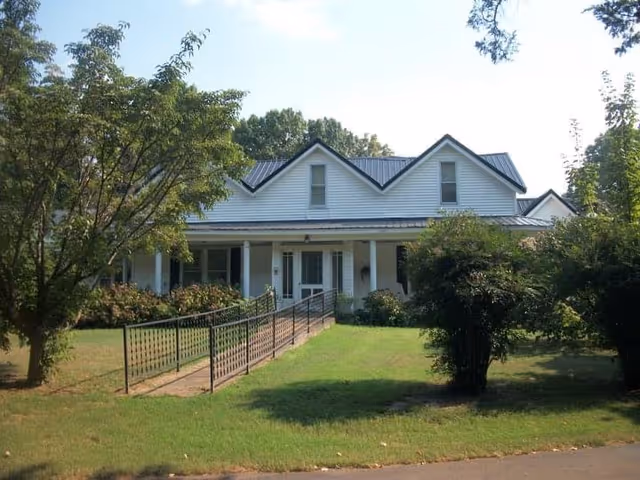 Ranch-style assisted living building with front porch, ramp, and surrounding trees.