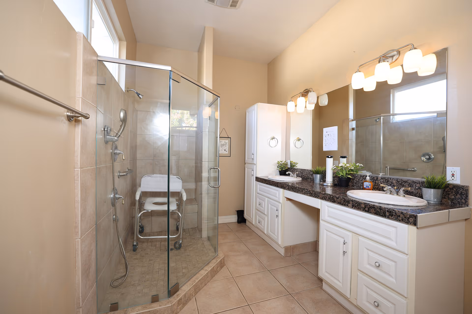 A spacious bathroom with beige tiled floors and walls, featuring a glass-enclosed shower with a white shower chair inside. There are two sinks with granite countertops, white cabinets underneath, and a large mirror above. The room is well-lit with two sets of wall-mounted light fixtures above the mirror. A window allows natural light into the space.