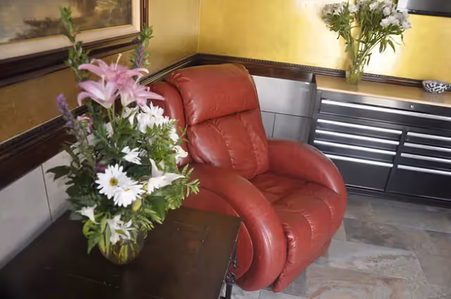 A cozy corner featuring a red leather recliner chair next to a dark wooden side table with a vase of mixed flowers including lilies and daisies. Behind the chair is a dark cabinet with drawers and another vase of white flowers on top, set against a yellow wall with a framed painting partially visible.