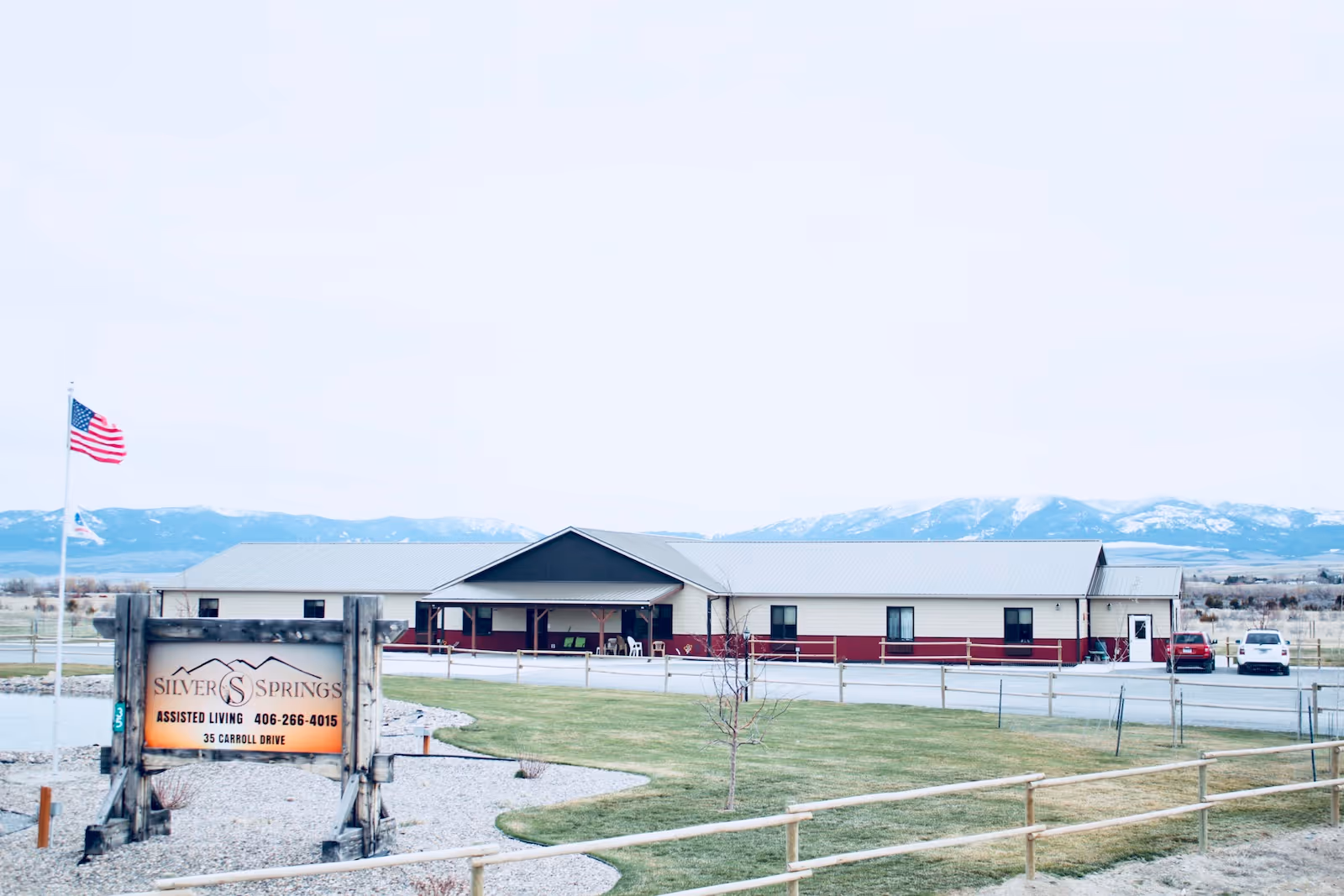 Exterior view of Silver Springs Assisted Living facility with a single-story building, a sign in the foreground displaying the facility name and contact information, an American flag on a flagpole, a fenced grassy area, and mountains in the background.