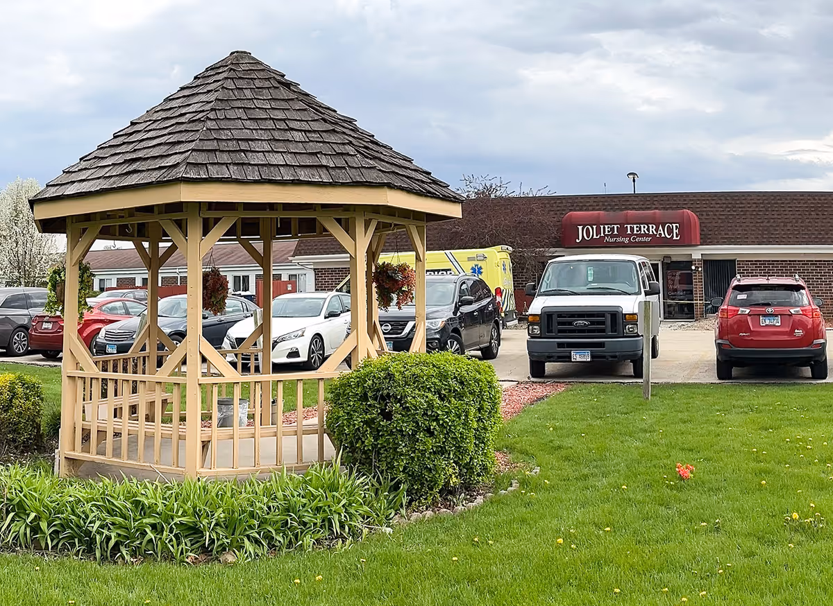 Outdoor view of Joliet Terrace Nursing Center showing a wooden gazebo in the foreground surrounded by green grass and plants. Several parked cars and a white van are visible in the parking lot in front of the building with a red awning displaying the facility name.