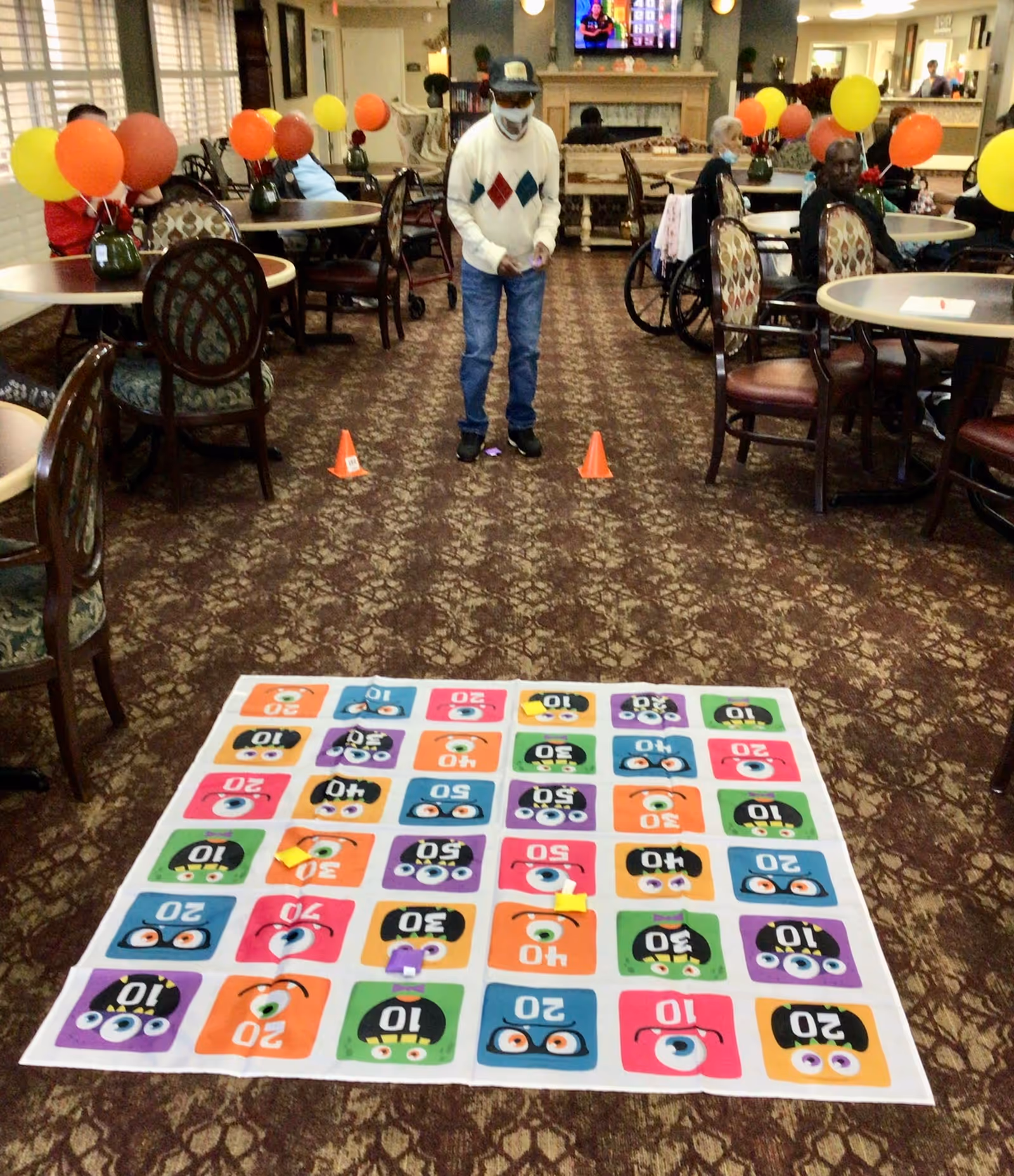 An elderly man wearing a mask and a sweater with diamond patterns is standing on a patterned carpet in a room with tables and chairs. In front of him is a colorful game mat with numbers and cartoon faces, and two small orange cones are placed on the carpet. Several other elderly people are seated at tables decorated with vases of balloons in yellow, orange, and red colors. The room has a cozy atmosphere with a fireplace and a TV mounted above it.