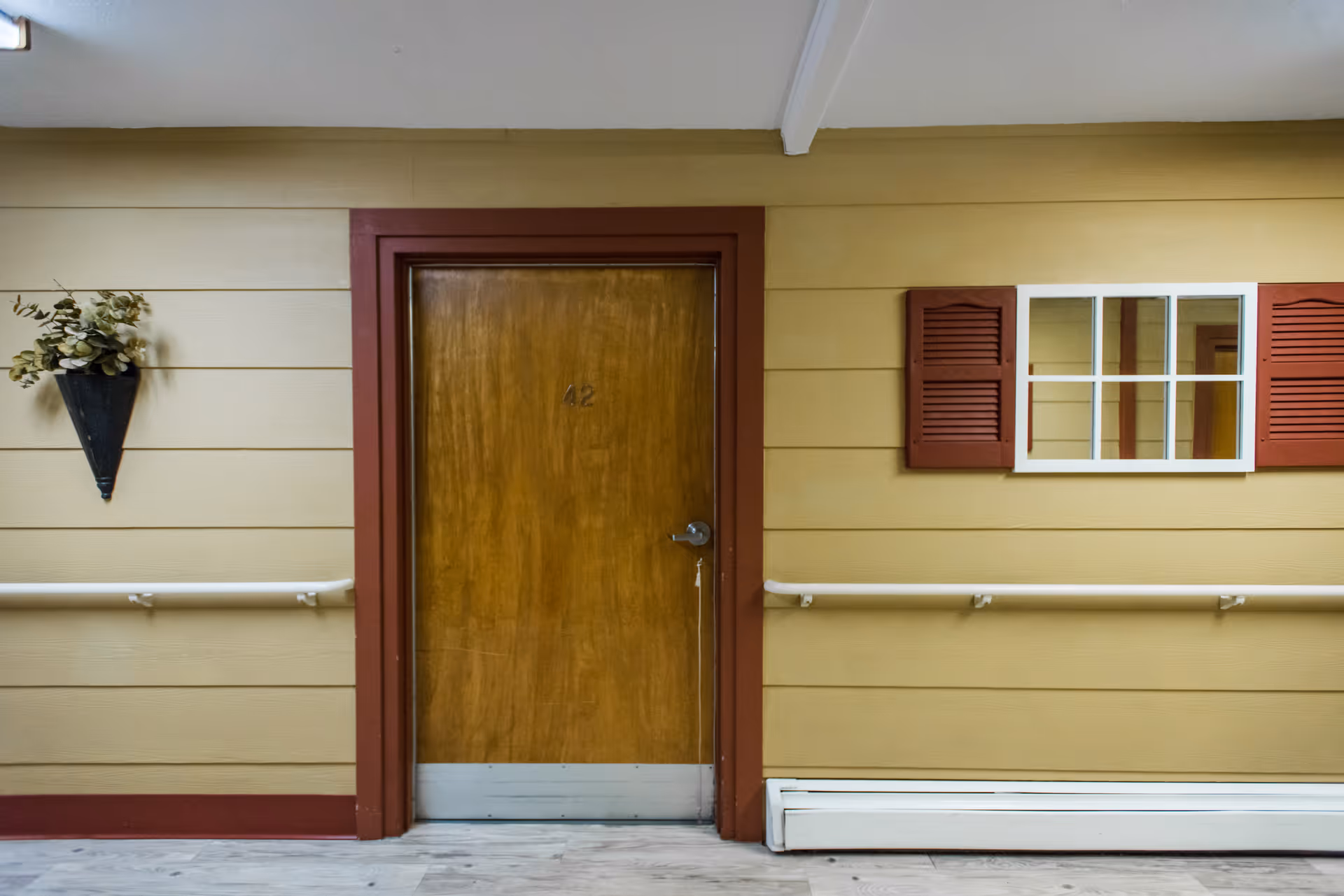 Interior hallway with a wooden door numbered 42, beige walls with horizontal paneling, a decorative wall planter with greenery on the left, a window with red shutters on the right, and white handrails along the wall.