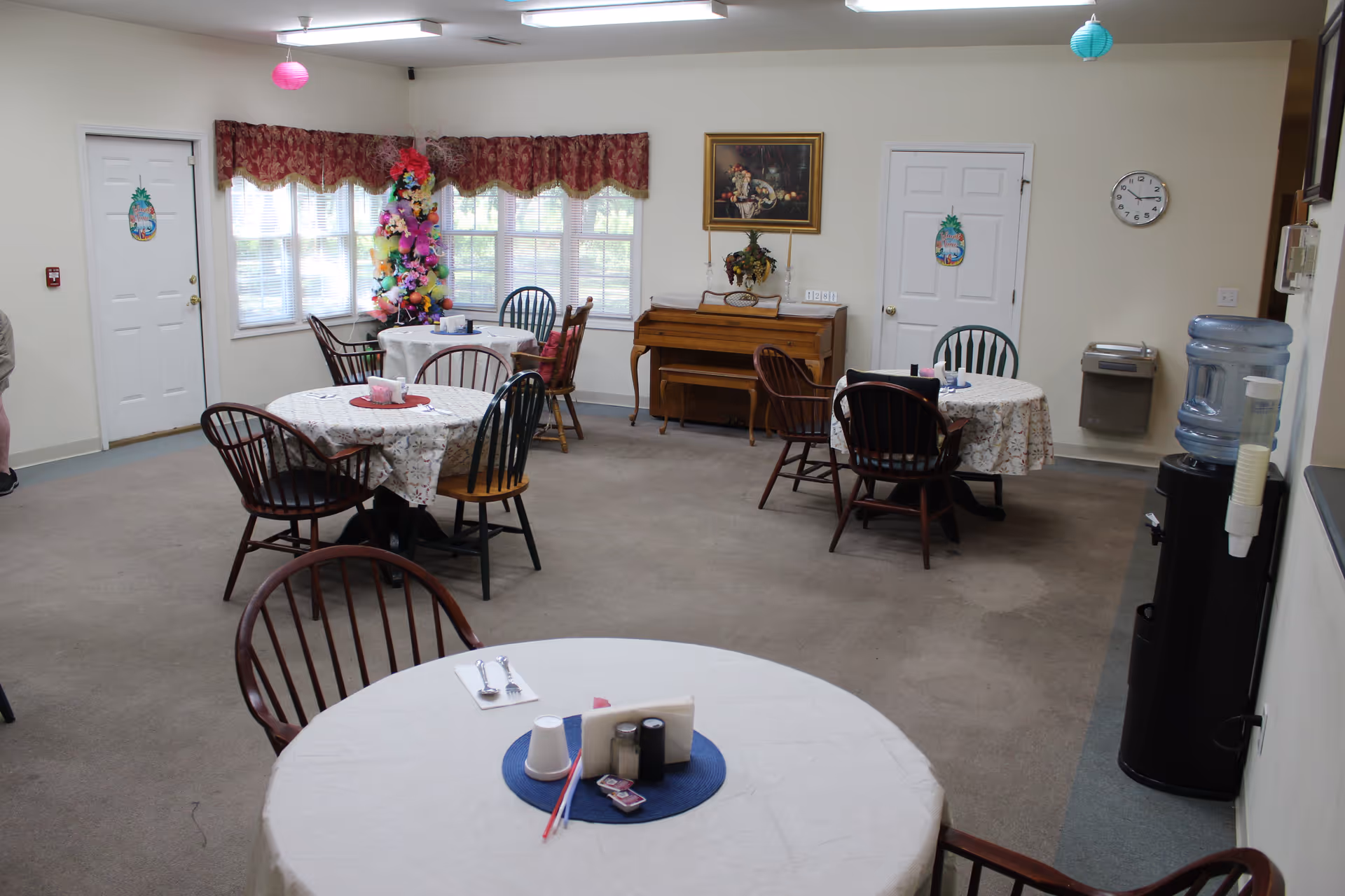 A dining room in Westbrook Assisted Living Facility with several round tables covered with white tablecloths and surrounded by wooden chairs. The room has large windows with red valances, a decorated Christmas tree in the corner, a piano against the wall, a water cooler, and a wall clock. The walls are light-colored and there are two white doors visible.