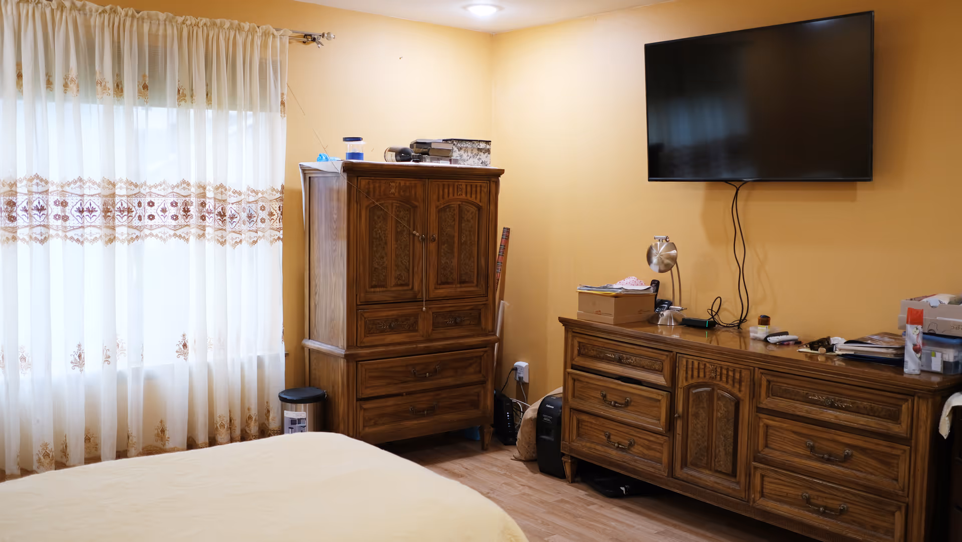 Bedroom in a senior home with a large wooden dresser, armoire, and wall-mounted TV.
