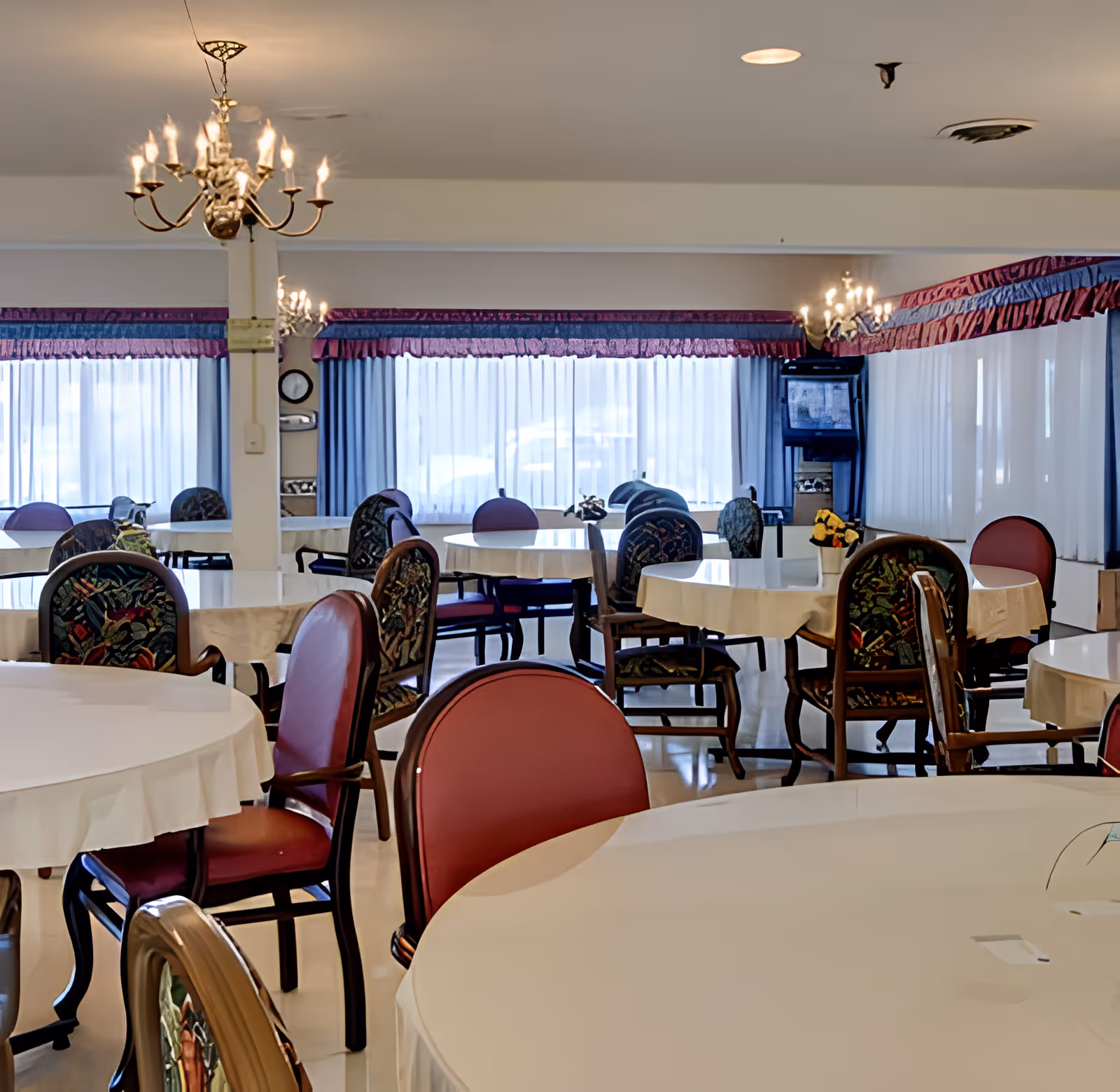 A spacious dining room with multiple round tables covered with white tablecloths and surrounded by chairs with red and patterned upholstery. The room features large windows with sheer white curtains and purple valances, and elegant chandeliers hanging from the ceiling providing warm lighting.