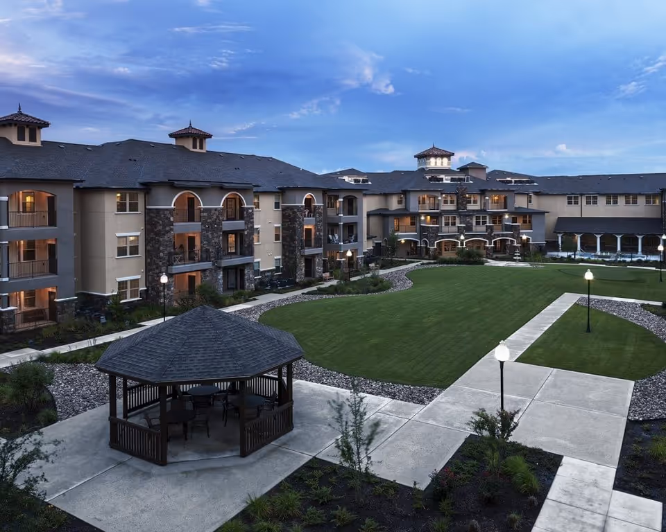 A landscaped courtyard with a gazebo, paved walkways, lampposts, and a multi-story senior living building in the background under a blue sky.