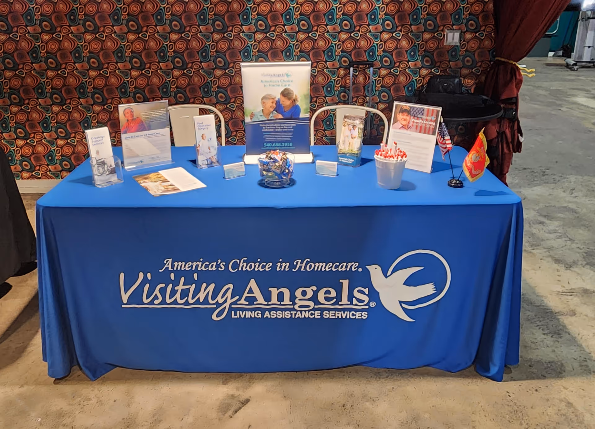 Indoor booth featuring a blue tablecloth, Visiting Angels banner, brochures, and small displays.