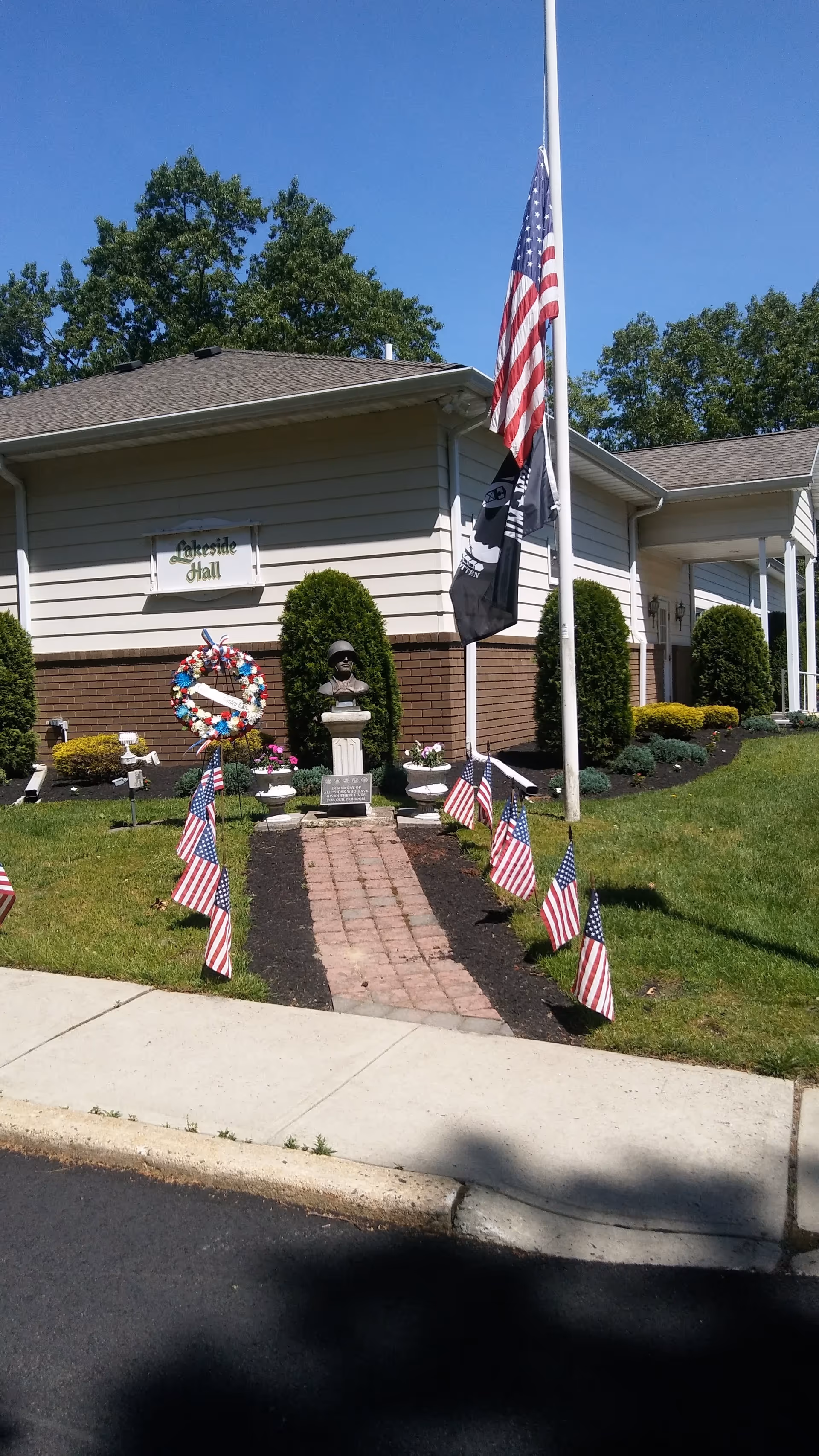 Facade of Cedar Glen Lakes exterior with flagpole, walkway, wreath, and American flags.