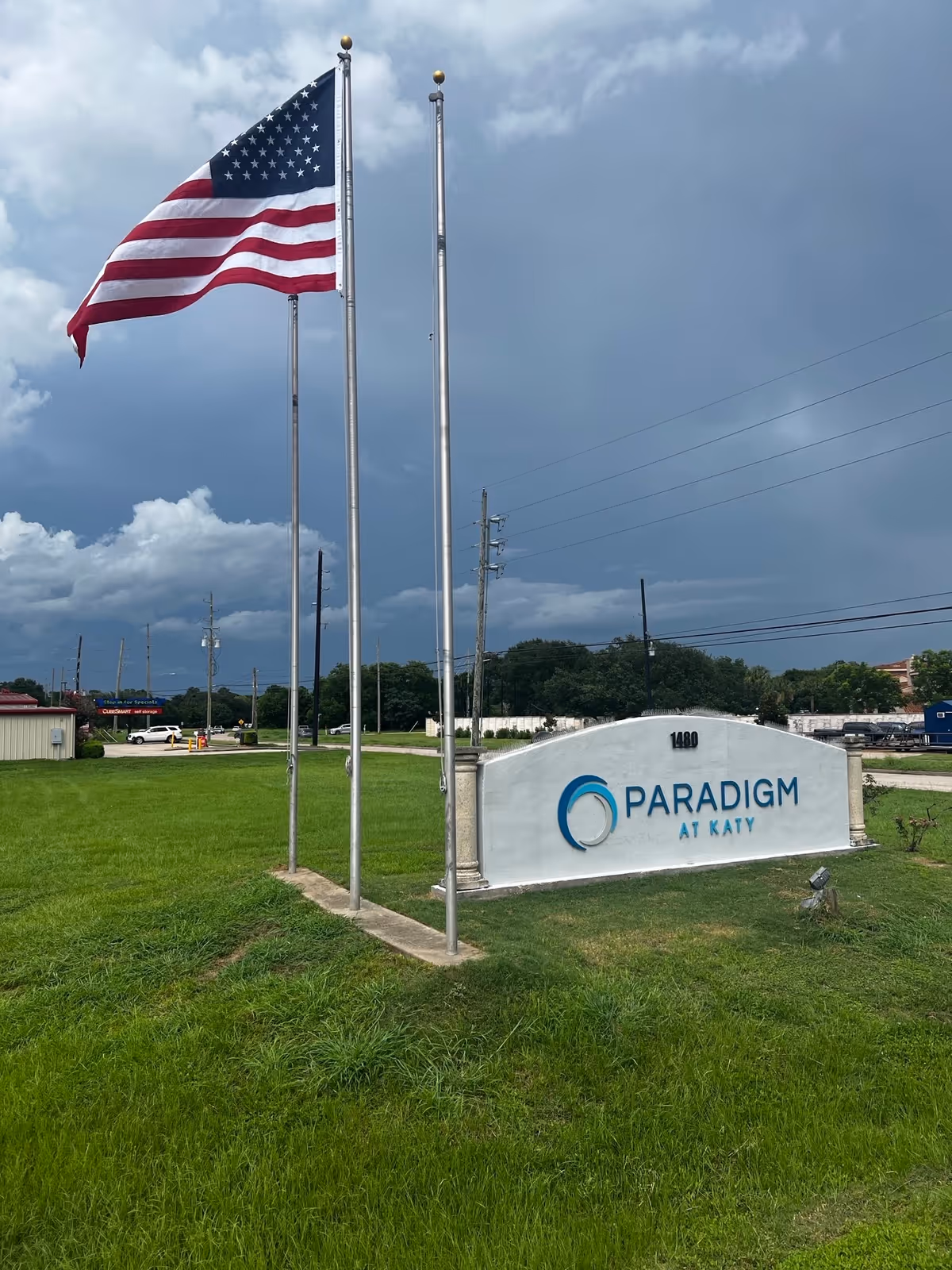 American flag on flagpoles beside a curved sign reading Paradigm at Katy on green lawn.