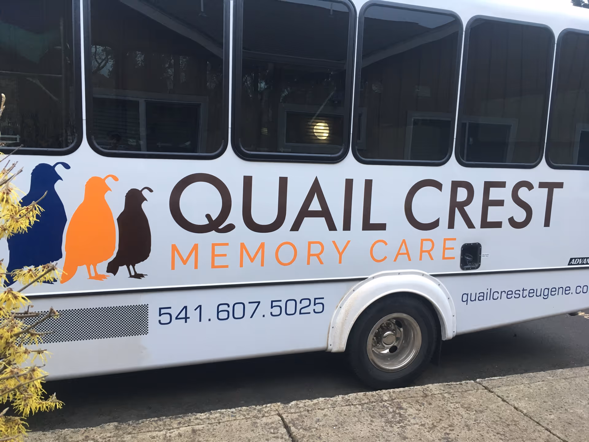 Side of a shuttle van displaying 'Quail Crest Memory Care' logo with three quail silhouettes and a phone number.