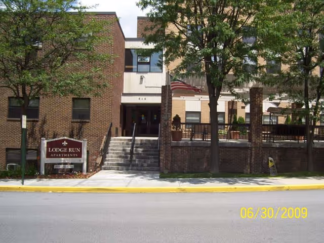 Front exterior of Lodge Run Apartments showing the entrance steps, a sign, and trees.