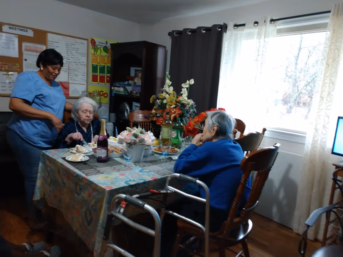 Two elderly women seated at a dining table with plates and flowers while a caregiver stands beside them in a bright dining room.