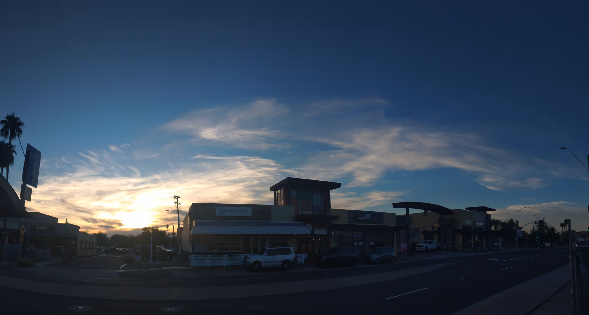 Exterior view of a shopping strip at sunset, with storefronts and parked cars along the street.