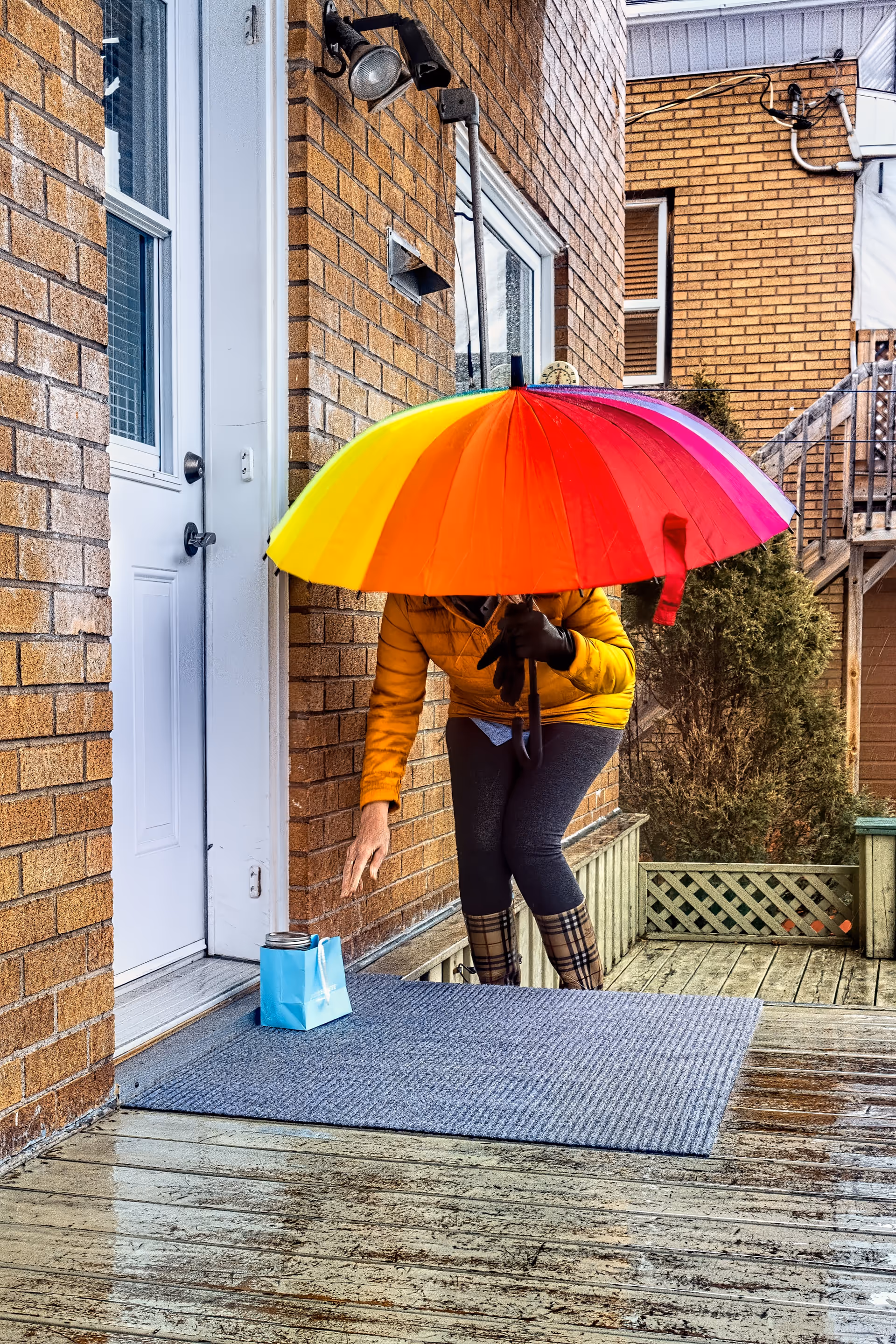 Person in yellow jacket under rainbow umbrella by front door on brick building.