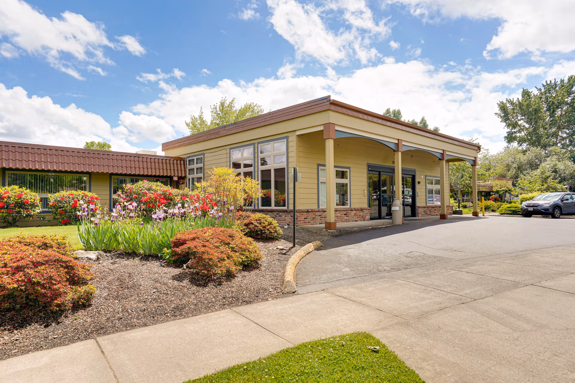 Exterior of Corvallis Manor Nursing and Rehabilitation Center with yellow siding, covered entrance, landscaping, and parking.