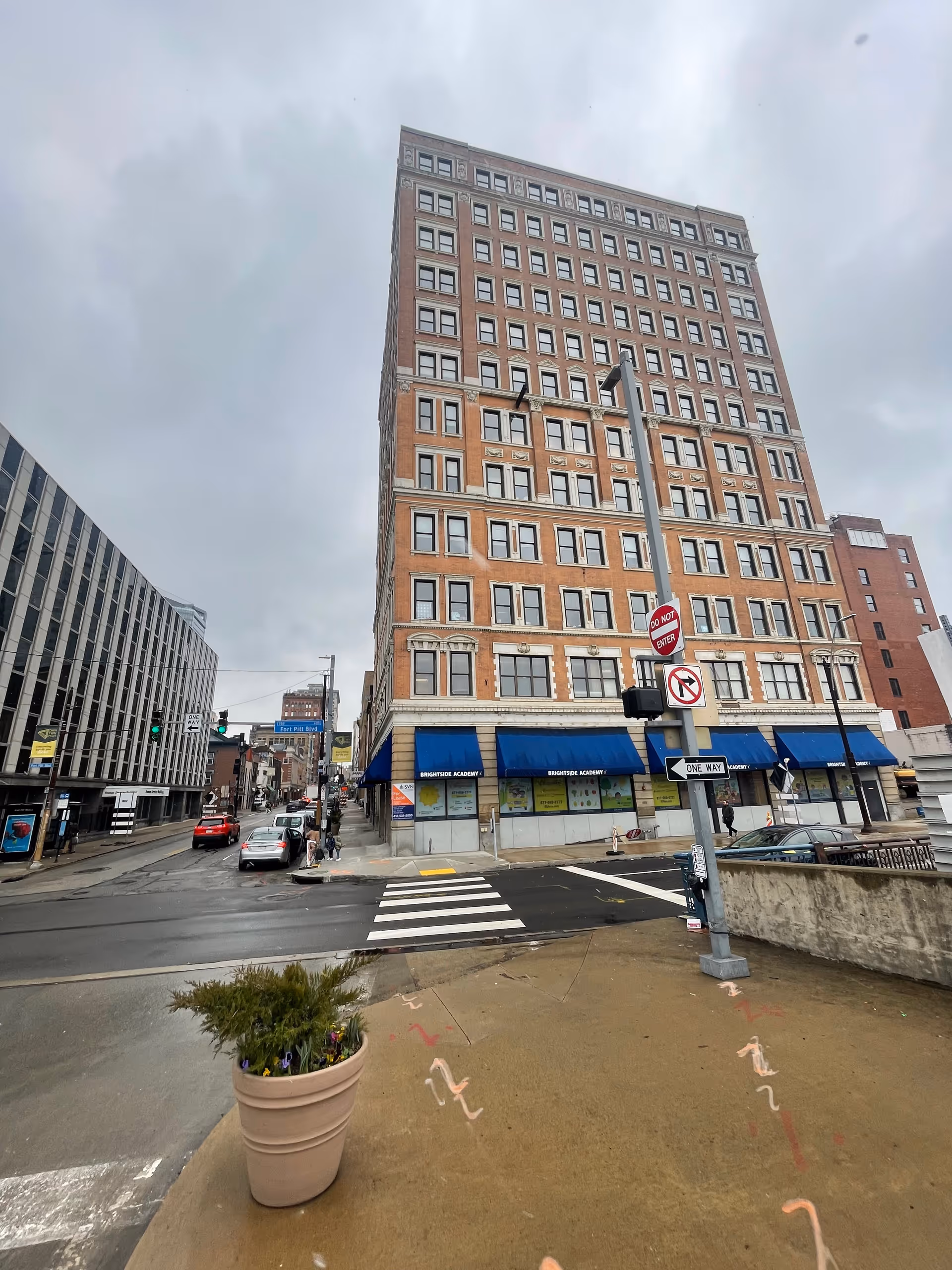 Tall brick building with blue awnings faces a busy city street and crosswalk.