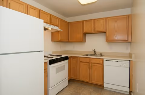 A small kitchen with wooden cabinets, a white refrigerator, a white electric stove with oven, a white dishwasher, a stainless steel sink, and beige countertops and walls.