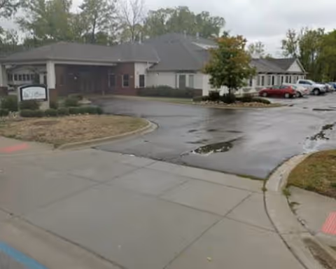 Exterior view of a senior living facility with a curved driveway and parked cars.