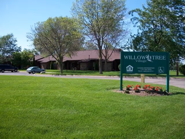 Exterior view of Willow Tree Village facility with a green lawn, several trees, a parked car, and a large green sign displaying the facility name and leasing information.