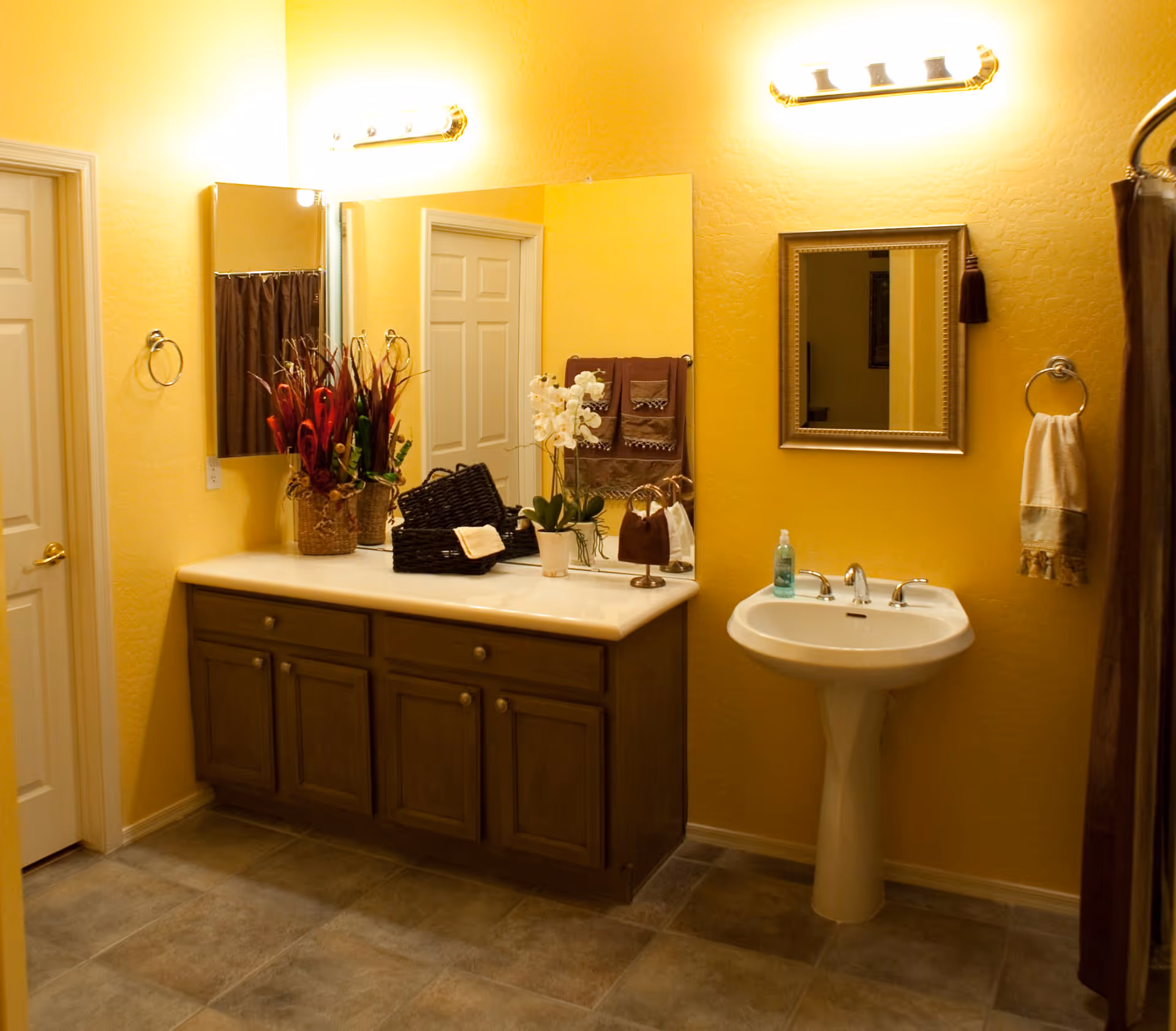 A bathroom with yellow walls and tiled floor featuring a wooden vanity with a large mirror, decorative plants, and baskets on the countertop. There is a white pedestal sink with a framed mirror above it, a soap dispenser, and towels hanging on the wall. Two light fixtures are mounted above the mirrors.