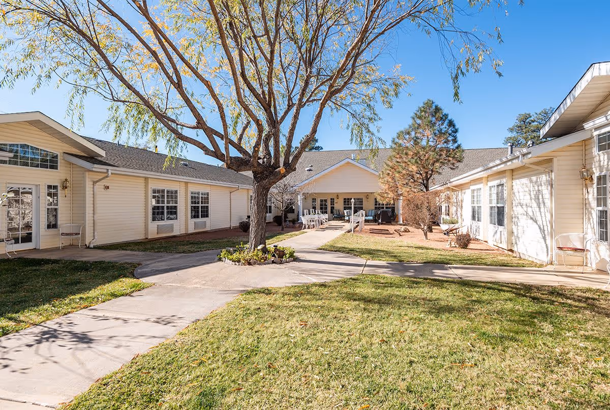 Outdoor courtyard area of Avista Senior Living Payson with a large tree in the center, surrounded by a paved walkway and green grass. The courtyard is enclosed by single-story beige buildings with multiple windows and doors, under a clear blue sky.