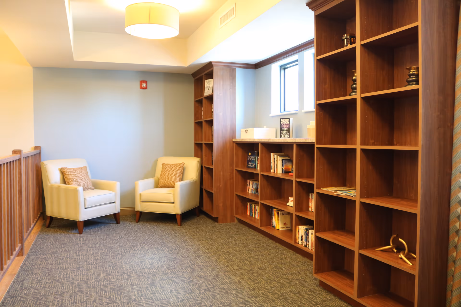 A cozy reading nook with two cream-colored armchairs each with a patterned cushion, positioned near a wooden railing. On the right side, there are large wooden bookshelves filled with books and decorative items. The room has a carpeted floor, light blue walls, and a ceiling light fixture.