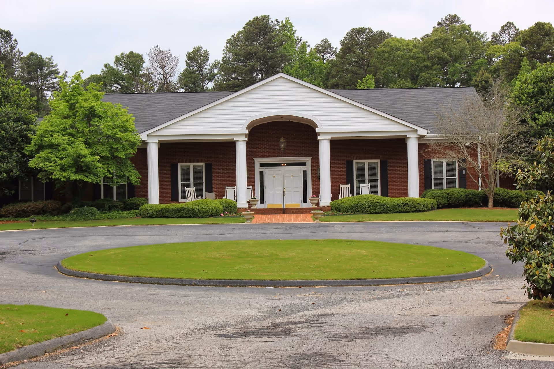 Front exterior view of a single-story brick building with white columns supporting a triangular pediment above the entrance. The entrance has double white doors and is flanked by windows with black shutters. There are rocking chairs on the porch and well-maintained bushes and trees surrounding the building. A circular driveway with a grassy center is in front of the building.