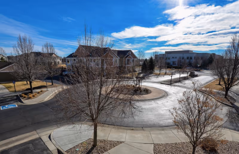 View of a roundabout driveway surrounded by leafless trees and buildings under a partly cloudy blue sky at Lakeshore at Centerra facility.