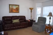 Cozy living room with leather sofa, recliner, lamps, painting, and sunny bay windows.