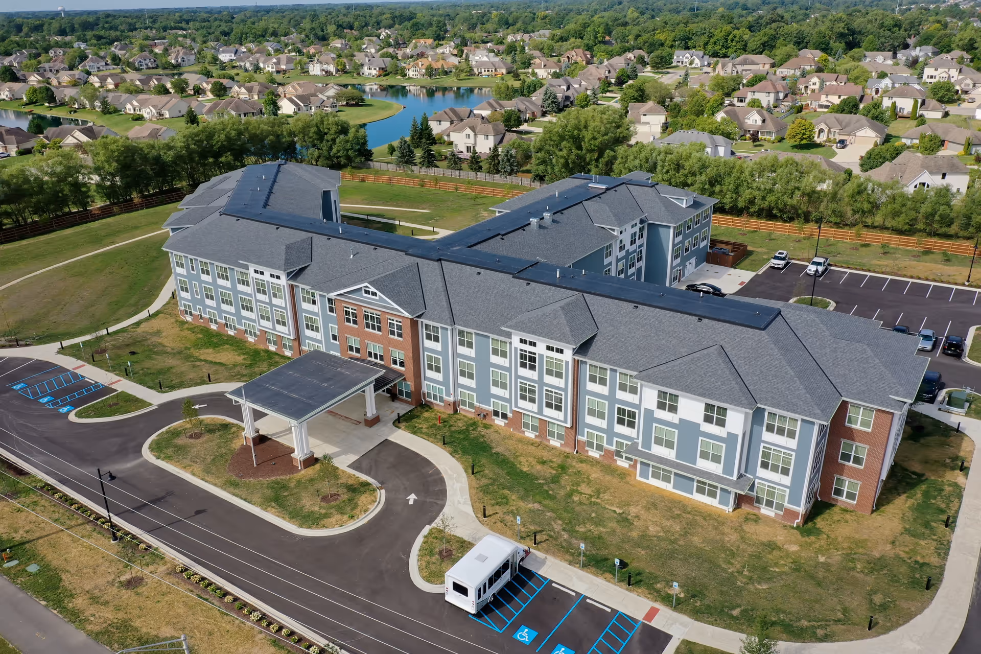 Aerial view of Evergreen Village at Fort Wayne, showing a large, modern three-story building with a covered entrance and surrounding parking lots. The building is surrounded by green lawns, trees, and a residential neighborhood with houses and a small pond in the background.