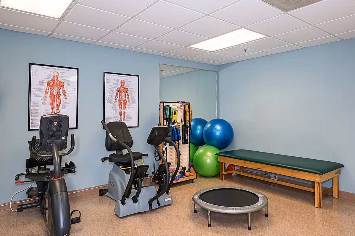 A small indoor gym/therapy room with exercise bikes, exercise balls, a trampoline, and a treatment table.