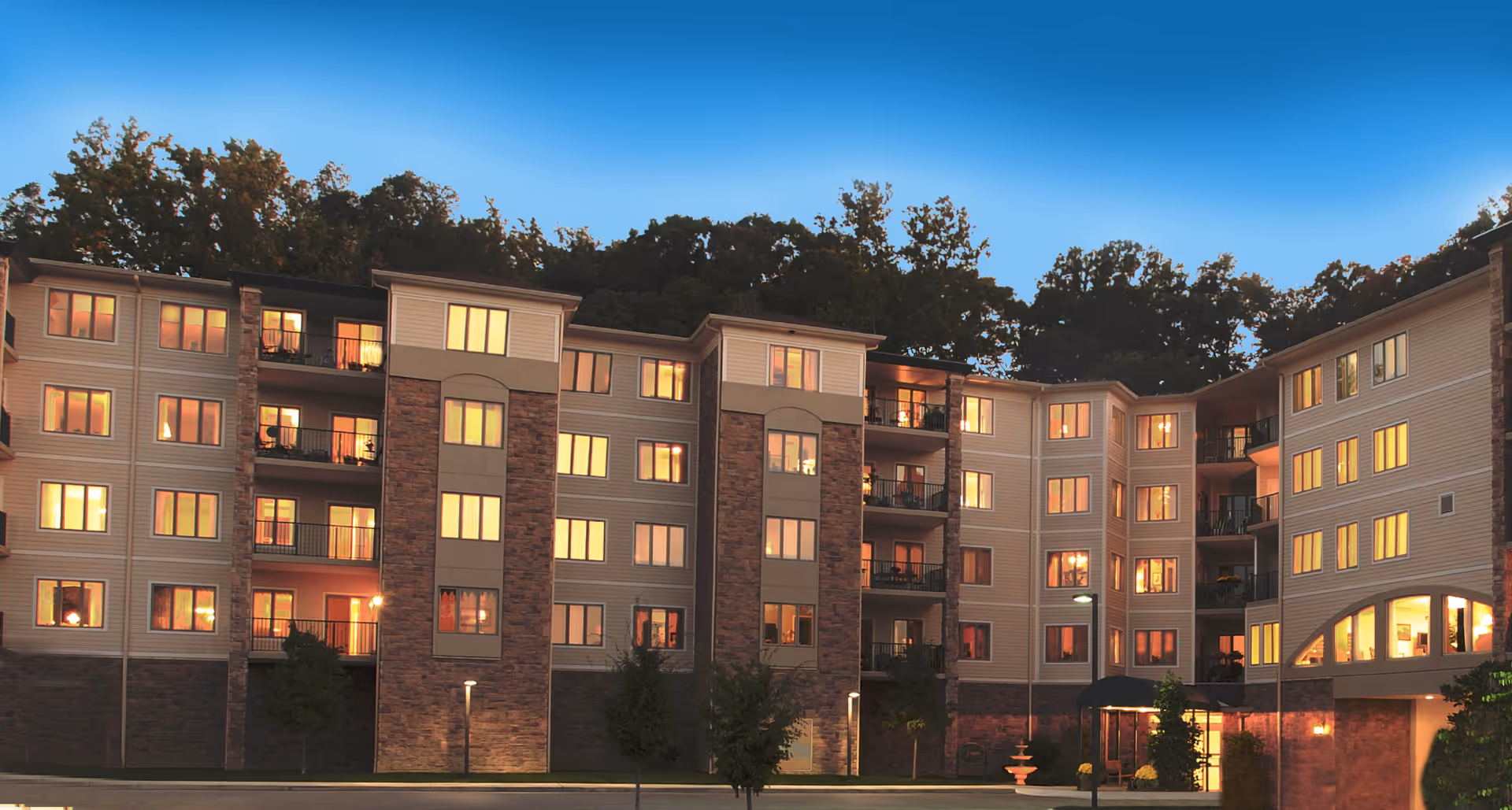 Exterior view of a multi-story senior living facility building at dusk with many windows illuminated from inside, surrounded by trees and a clear sky.