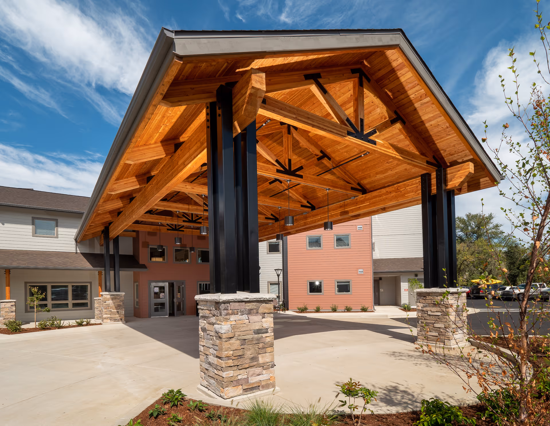 Entrance area of The Landing a Senior Living Community featuring a large wooden canopy supported by black metal and stone pillars, with the building facade and parking lot visible in the background under a partly cloudy sky.
