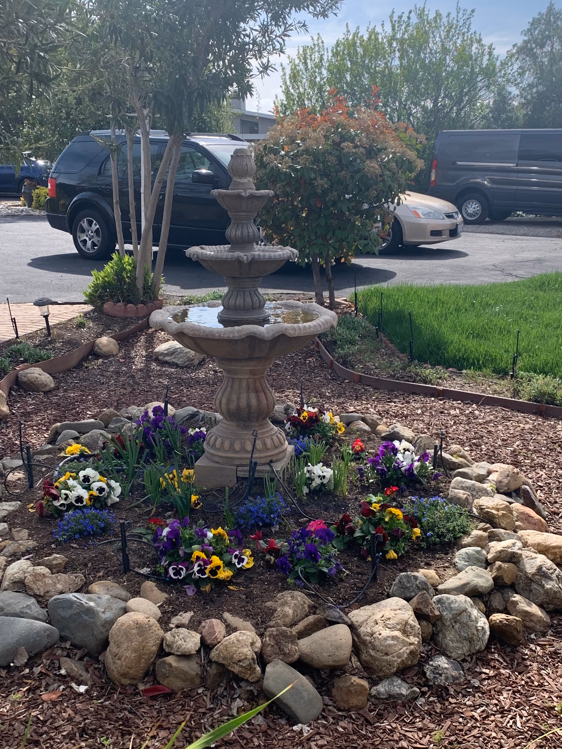 Stone tiered fountain surrounded by colorful flowers in a landscaped garden area.