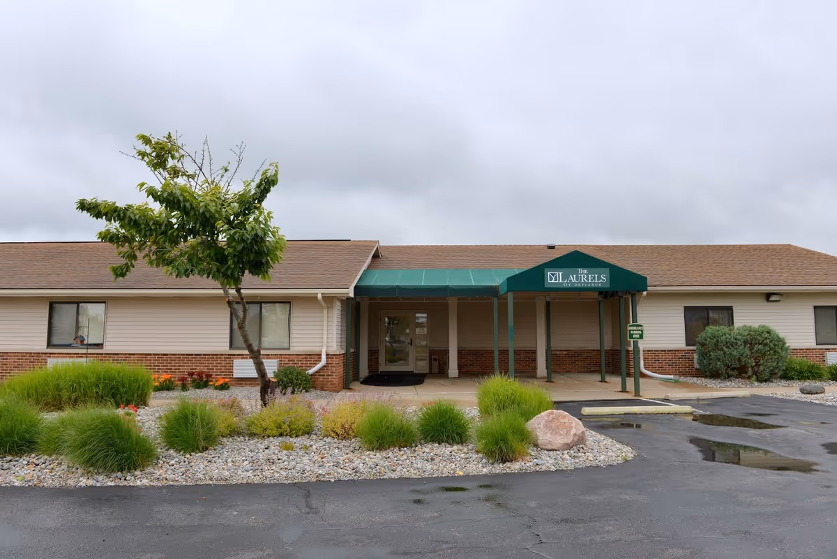 Exterior view of The Laurels of Defiance building with a green awning over the entrance, surrounded by landscaping with bushes, a small tree, and rocks. The sky is cloudy and the parking lot in front has some puddles.