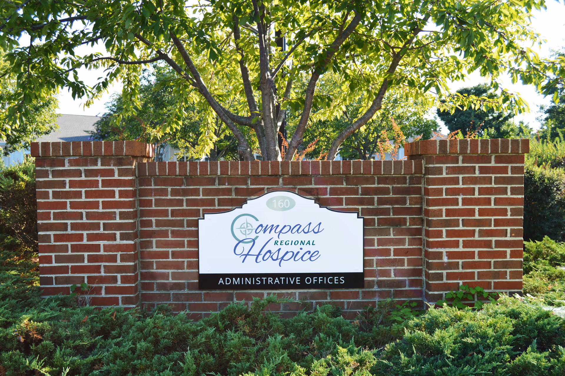 Brick entrance sign for Compass Hospice Administrative Offices, with greenery and trees behind.