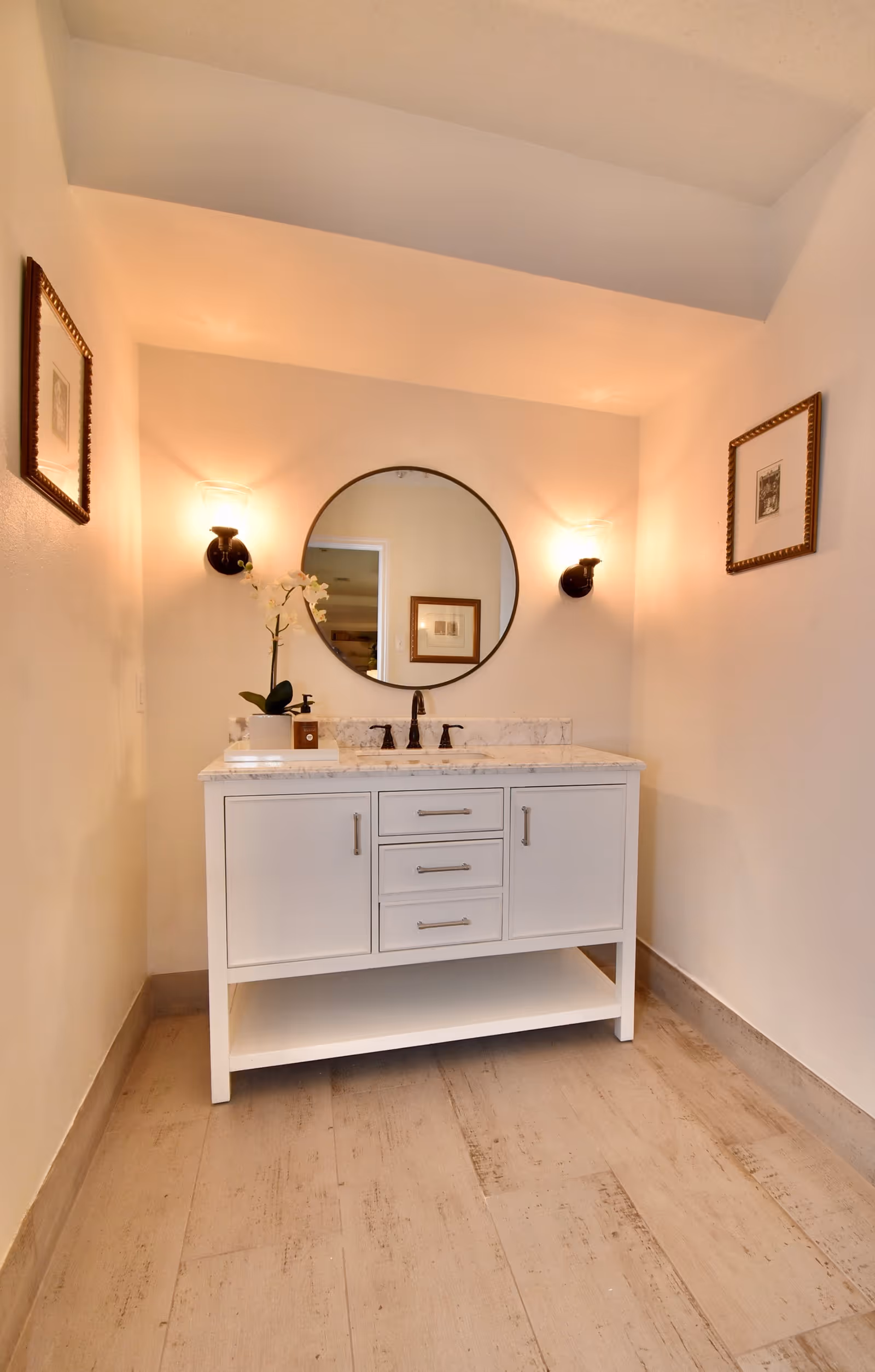 A clean and modern bathroom vanity with a white cabinet and marble countertop. Above the vanity is a round mirror flanked by two wall-mounted light fixtures. There is a small potted orchid plant on the countertop. The walls are painted a light color and two framed pictures hang on either side of the vanity.