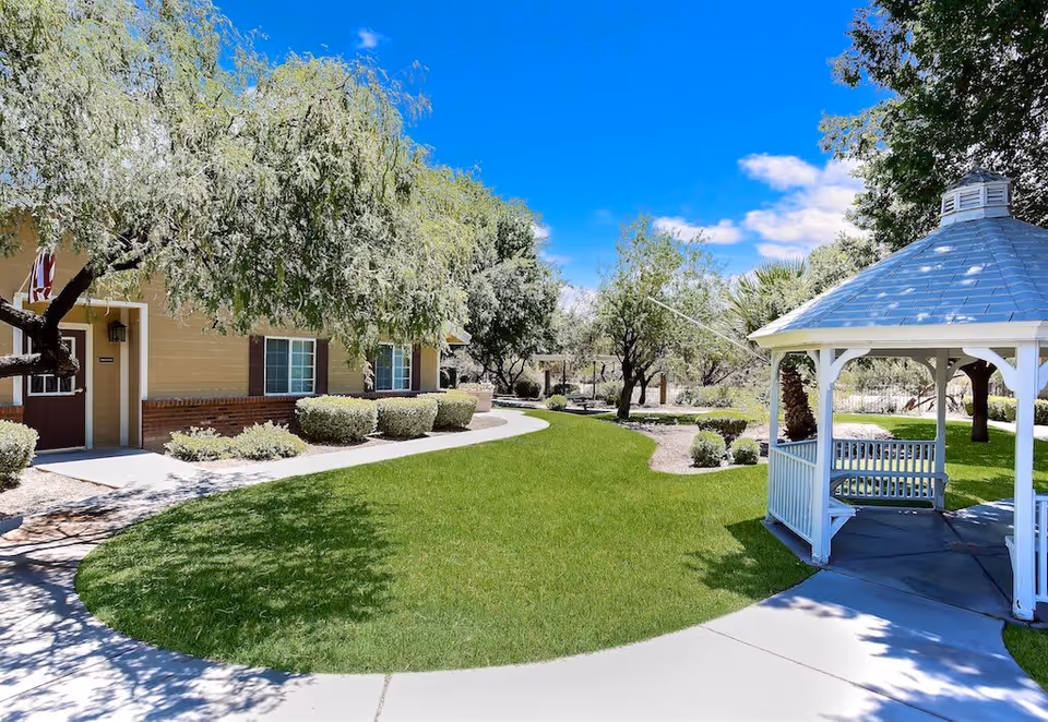 Outdoor garden area at Tucson Gardens Memory Care featuring a well-maintained green lawn, a white gazebo with a shingled roof, trees, shrubs, and a beige building with windows and a door in the background under a clear blue sky.