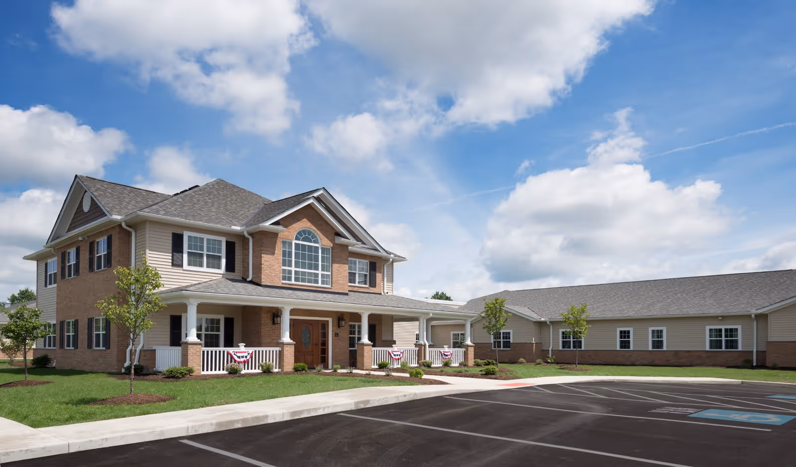 Exterior view of a two-story senior living facility building with brick and beige siding, a covered porch decorated with patriotic bunting, small trees and shrubs in the landscaped lawn, and an empty parking lot under a partly cloudy blue sky.