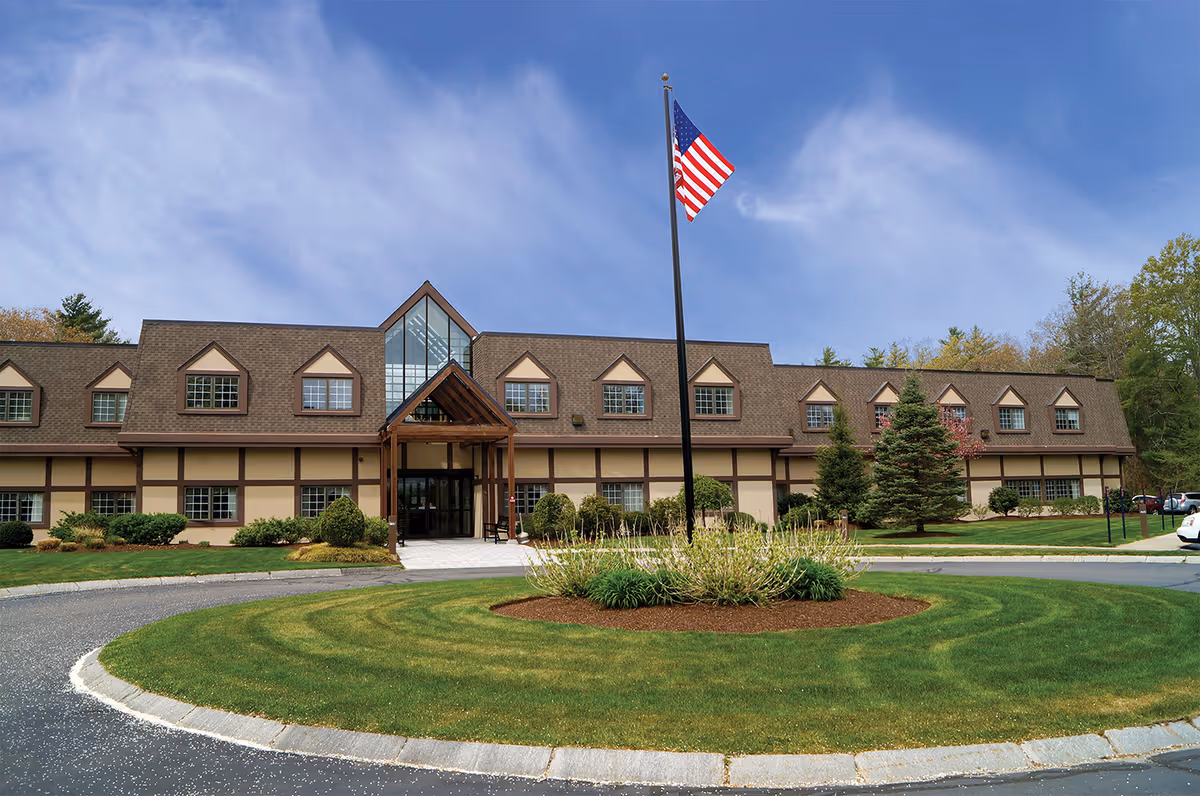 Front exterior view of Sippican Health Care Center Inc, a two-story building with a brown roof and beige walls, featuring multiple windows and a central entrance with a glass facade. In front of the building is a circular driveway with a landscaped island in the center, which has an American flag on a tall flagpole. The sky is clear with some clouds.