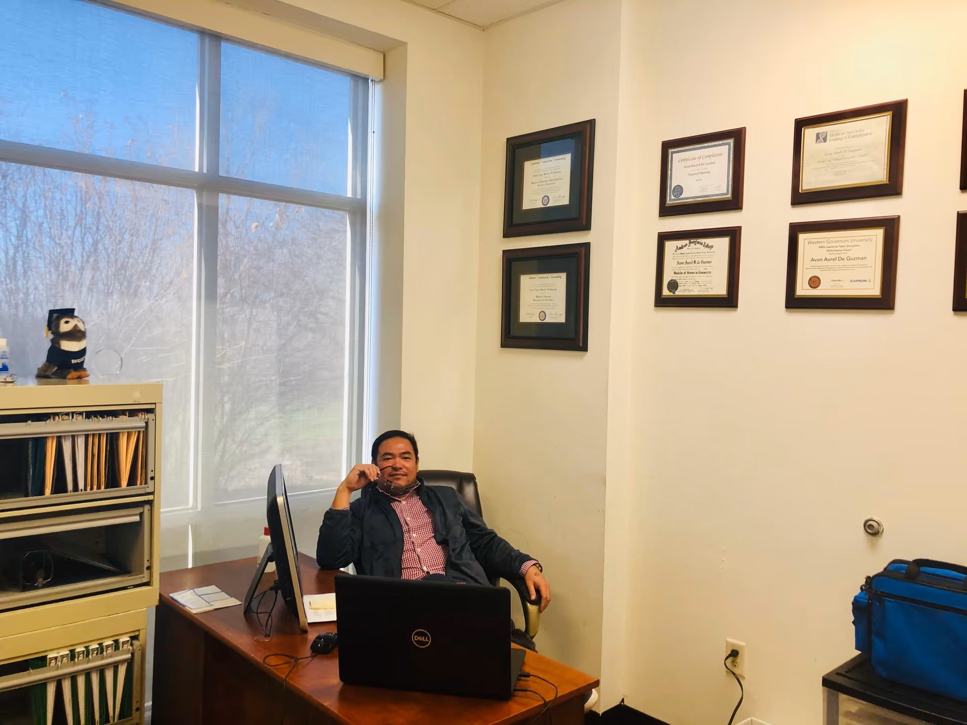 Office scene with a man at a desk, computer, and framed certificates on the wall.