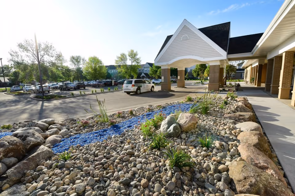 Front entrance and porte-cochere of a senior living facility with rocky landscaping, blue decorative stones, and parked cars in the lot.