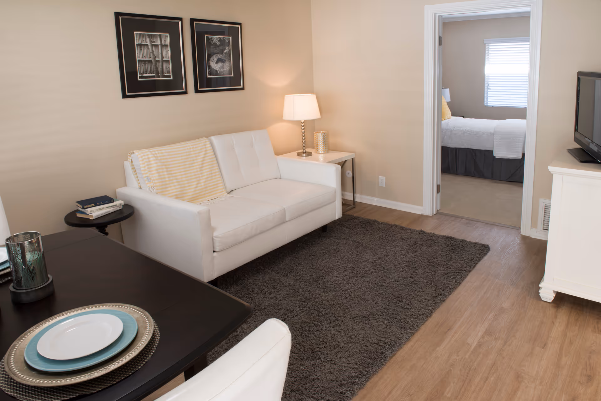 Bright living room with white sofa, rug, dining table, and doorway to a bedroom.
