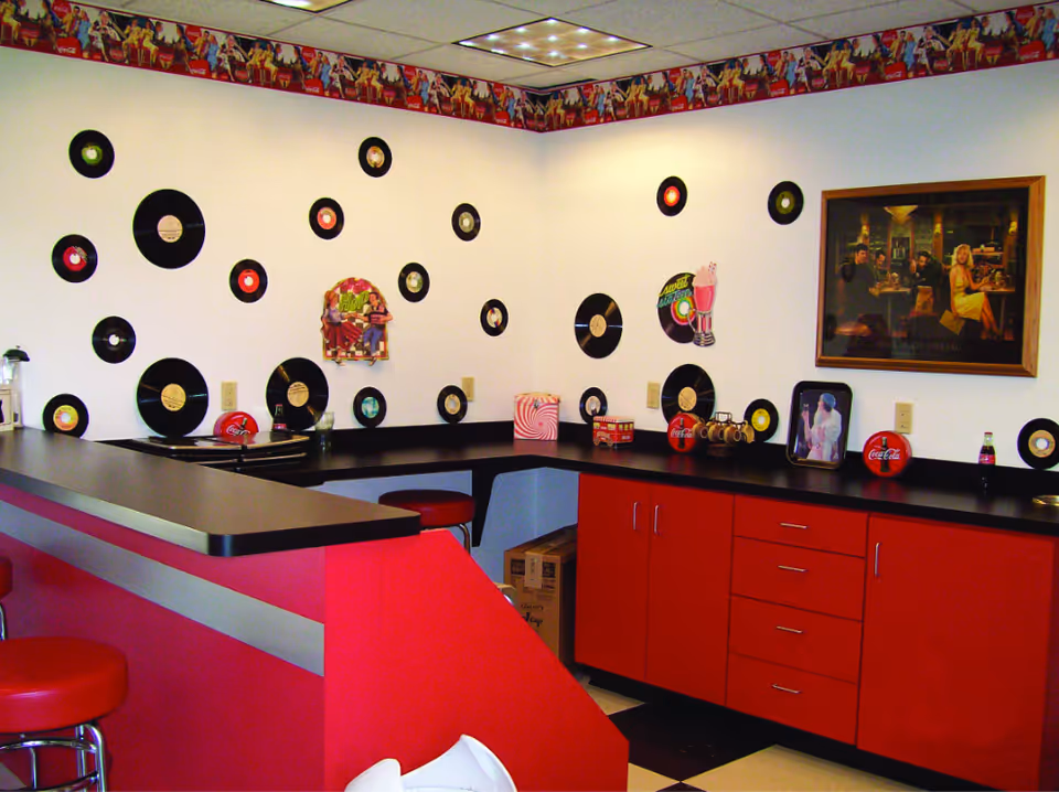 Retro soda-shop style kitchenette with red cabinets and barstools, black countertops, and vinyl records decorating the white walls.
